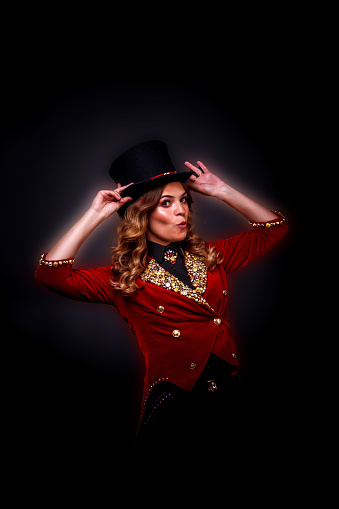 Woman magician illusionist circus in theatrical clothes showing and smiles at black background. Female actress in stage costume and top hat on head. Concept of performance. Copy text space