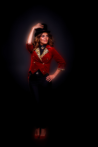 Female magician illusionist circus in theatrical clothes showing and smiles at black background. Young woman actress in stage costume and top hat on head. Concept of performance. Copy text space