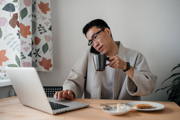 istock Middle aged asian man in spectacles with smartphone and laptop in kitchen. 1421313863