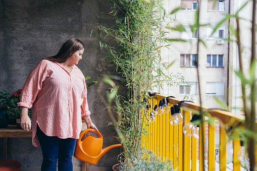 Plus size Caucasian woman watering her houseplants on the balcony with an orange watering can.