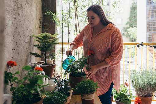 Caucasian woman taking care of her potted plants on the balcony. She is watering them using a blue jug.
