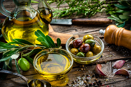 Close up view of  two glass bowls filled with olives and extra virgin olive oil shot on rustic wooden table. Olive tree branches, garlic, salt and peppercorns complete the composition. High resolution 42Mp studio digital capture taken with Sony A7rII and Sony FE 90mm f2.8 macro G OSS lens