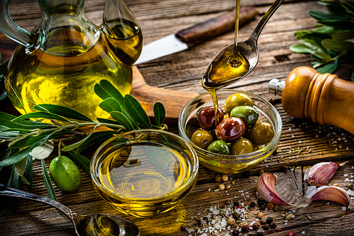 Front view of an olive oil bottle surrounded by green olives and a olive wooden cutting board with more green olives, olive branches and a withe bowl filled with three green olives and olive oil on top. Low key DSLR photo taken with Canon EOS 6D Mark II and Canon EF 24-105 mm f/4L