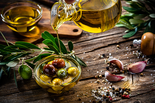 Close up view of  an extra virgin olive oil bottle pouring oil into a glass bowl filled with a variety of organic olives. Olive tree branches, garlic, salt and peppercorns complete the composition. High resolution 42Mp studio digital capture taken with Sony A7rII and Sony FE 90mm f2.8 macro G OSS lens