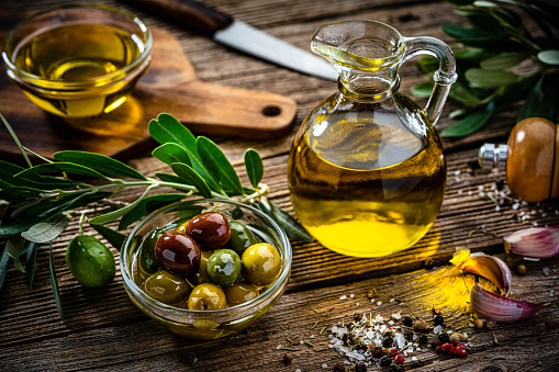 Close up view of an extra virgin olive oil bottle and a glass bowl filled with various types of organic olives shot on rustic wooden table. Olive tree branches, garlic, salt and peppercorns complete the composition. High resolution 42Mp studio digital capture taken with Sony A7rII and Sony FE 90mm f2.8 macro G OSS lens