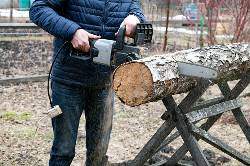 A man in jeans saws a birch log with an electric saw outdoors. Preparing firewood for the winter