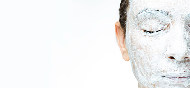 Caucasian female is having a white facial mask in his face. Even on her eyes and lips in front of white background. Her eyes are closed.