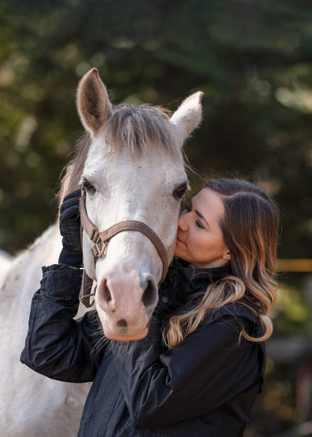 young woman standing next to white arabian horse eyes closed as if she's kissing or smelling, blurred trees background, closeup detail - horse arabian horse arabia white imagens e fotografias de stock