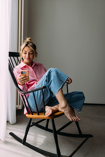 A smiling beautiful casually dressed Caucasian entrepreneur holding her smartphone and watching something online while sitting in her cozy chair.