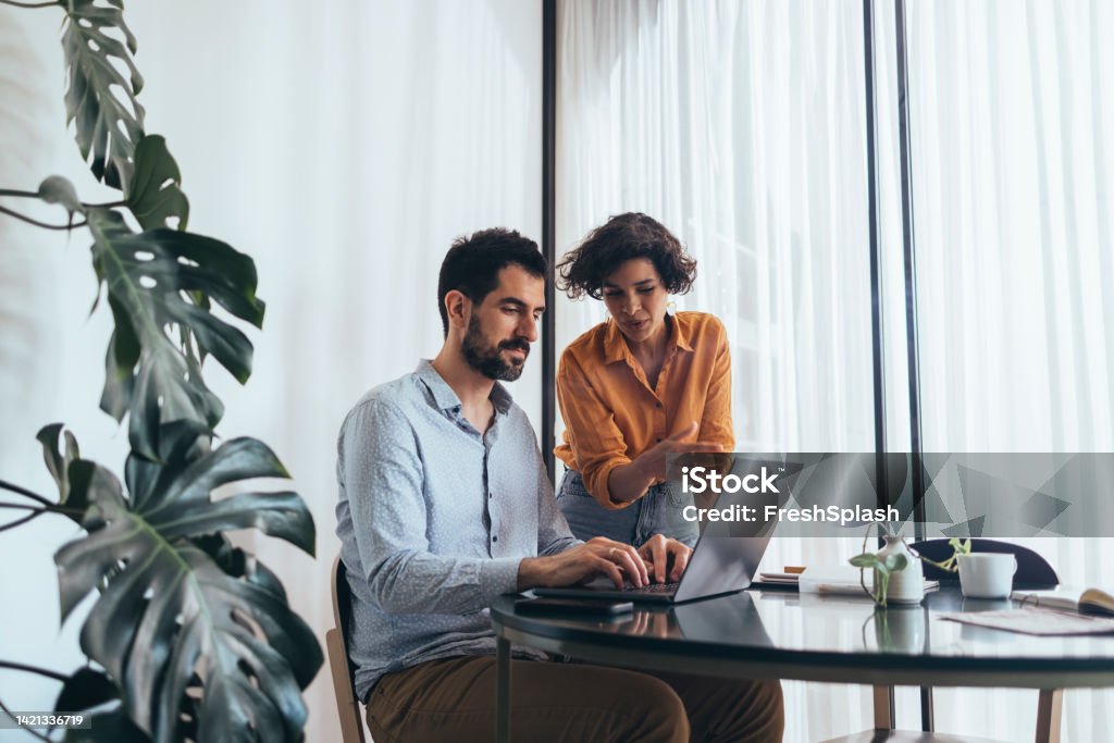 Two Colleagues Cooperating  At Work A smiling Latin-American businesswoman helping her Caucasian colleague at the computer while he is typing something. They are in the office. Mentorship Stock Photo