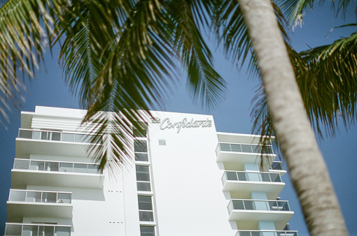 Miami Beach, Florida; March 2020; Art Deco details of The Confidante Hotel, Miami Beach, Florida with white exterior, and light blue accents, as viewed from the beach with palm tree accent.