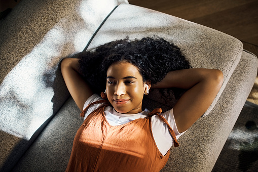 Relax woman with earphones listening to podcast or music on sofa with sunshine. Gen z black girl or lazy teenager enjoying playlist, audio or songs on wireless technology on a couch on the weekend