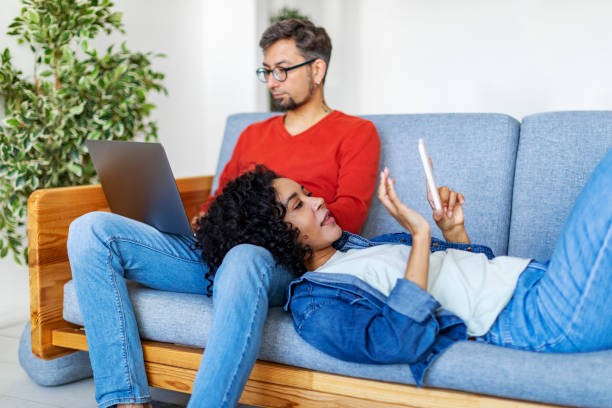 multiracial couple using laptop and mobile at home for work and leisure - multi ethnic group concentration mobile phone using laptop imagens e fotografias de stock