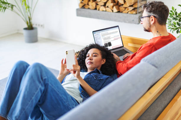 multiracial couple using laptop and mobile at home for work and leisure - multi ethnic group concentration mobile phone using laptop imagens e fotografias de stock