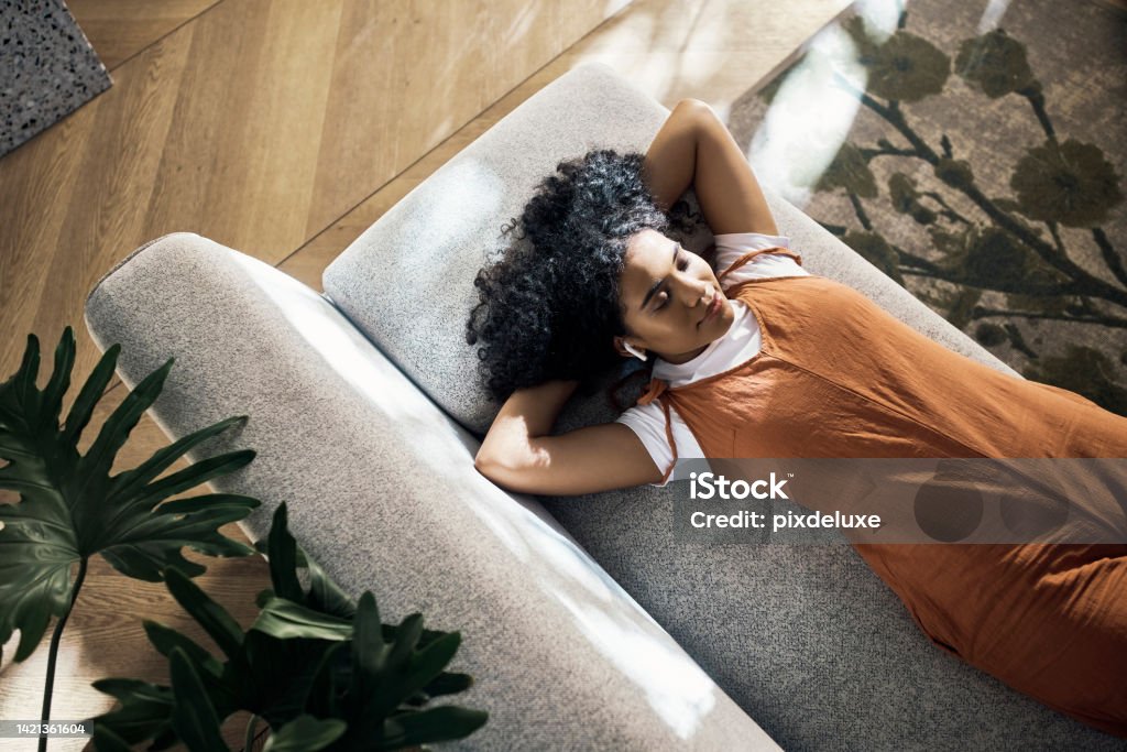 Sofa, girl and wireless earphones with music to relax with comfortable lying down position rest. Woman streaming soothing app in living room for tranquil and calm chill break time alone. Sofa, girl and wireless earphones to relax with comfortable  lying down position for chill break. Woman listening to soothing music or podcast in living room for tranquil and calm time alone. Music Streaming Service Stock Photo