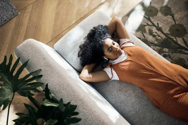 Sofa, girl and wireless earphones to relax with comfortable  lying down position for chill break. Woman listening to soothing music or podcast in living room for tranquil and calm time alone.