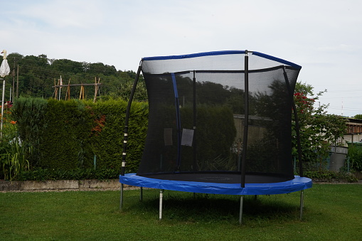 Empty blue trampoline with safety net on the lawn in garden. In the background is green vegetation creating hedge.