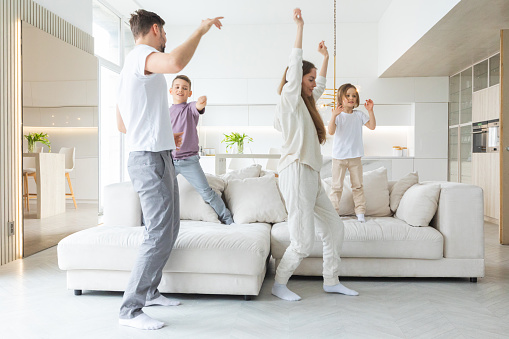Funny active family of four young adult parents and cute small children dancing together in living room interior, carefree little kids with mum dad having fun laughing enjoy leisure at home