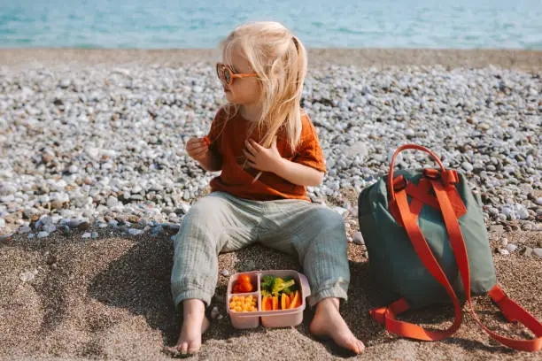 Child eating vegetables with lunchbox outdoor travel vacations healthy lifestyle vegan food picnic on beach girl toddler with lunch box and backpack
