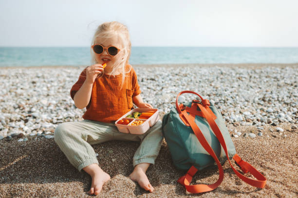 Child with lunch box eating vegan food outdoor travel summer vacations picnic on beach healthy lifestyle toddler girl with lunchbox snacks and backpack stock photo