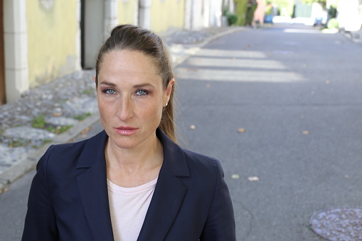 A serious businesswoman is posing with confidence in urban environment. She has a pony tail, wears a suit and has blue eyes