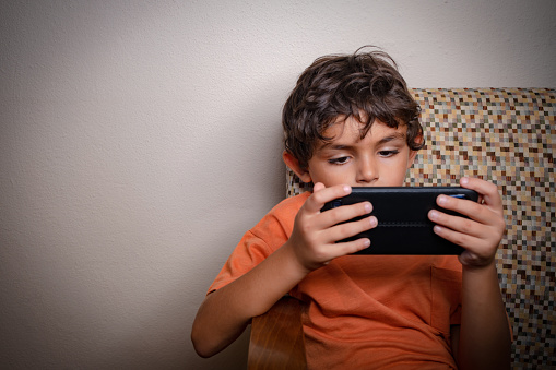 6 years old boy, brunette, with messy hair. He is sitting on the sofa inside the house. He has a black smartphone in his hand. The boy is wearing an orange T-shirt.The child is staring at the screen of the phone he is holding horizontally with all his attention.Portrait shooting.Space for text.