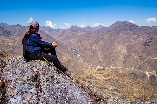 Latin woman, sitting on a large rock, contemplating the Peruvian Andes
