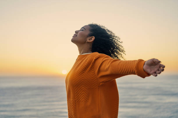 libertà, donna felice e rilassante in spiaggia che si allunga con le braccia aperte durante il tramonto. le donne si sentono felici e si godono un'ora d'oro nella natura con un cielo colorato e uno sfondo oceanico in estate - young adult women african descent 30s foto e immagini stock