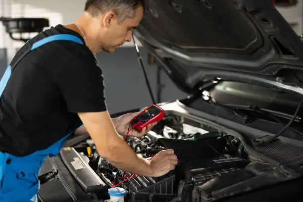 Photo of Professional mechanic is working in car service. Good, electrician, electricity, battery charged fully. Check battery voltage with electric multimeter. Car starter battery