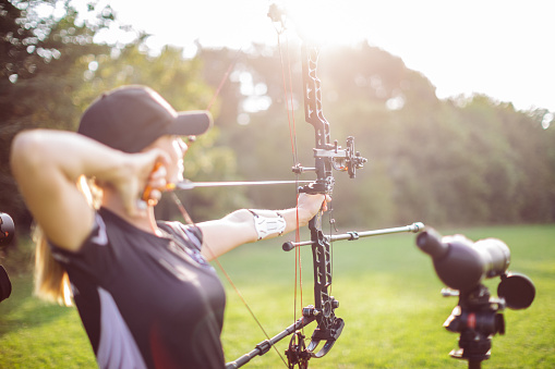 Woman practicing archery training with compound bow on open field before sunset. She is in very good shape and well equipped. Focus is on the bow