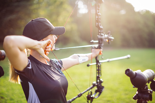 Woman practicing archery training with compound bow on open field before sunset. She is in very good shape and well equipped. Focus is on the woman