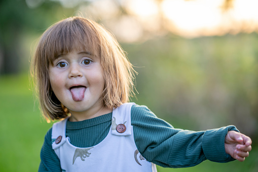 A sweet little toddler of Hawaiian decent, stands outside as she looks to the camera with a smile.  She is dressed comfortably in a jumper and is sticking out her tongue as she is silly for the camera and enjoys the fresh air.