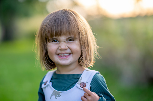 A sweet little toddler of Hawaiian decent, stands outside as she looks to the camera with a smile.  She is dressed comfortably in a jumper and has a big smile on her face as she enjoys the fresh air.