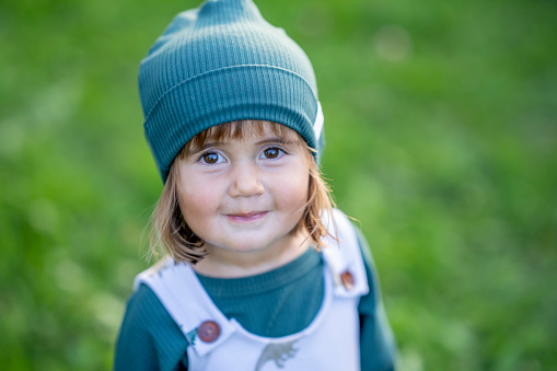 A sweet little toddler of Hawaiian decent, stands outside as she looks to the camera with a smile.  She is dressed comfortably in a jumper and has a hat on as she enjoys the fresh air.