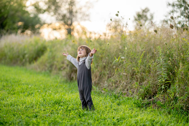 A sweet little brunette haired girl of Hawaiian decent, stands outside in the grass as she soaks in the sunlight and nature.  She is dressed casually in a jumper and has her hands stretched out as she looks up to the sky.