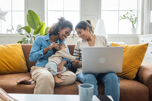 Young mothers with baby girl in living room. One woman using laptop for work or online shopping.