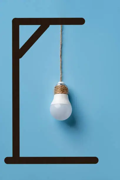 Light bulb hanging from gallows over blue background. Deindustrialization and regression. Reduction of manufacturing, removal of industrial activity concept. Economic crisis. Vertical frame.