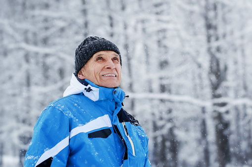 Portrait of mature grandfather who walking at winter forest and looking with smile. Healthy lifestyle of senior human.
