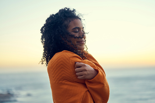 Comfort, winter and freedom with black woman by the ocean seaside alone in cold nature. Hug, embrace and relax with young female with a jersey by the beach on calm, peaceful holiday