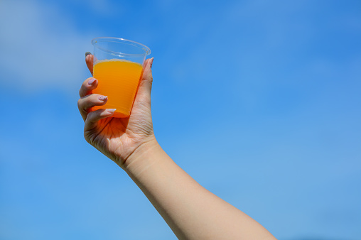 Image of woman hand holding orange juice in disposable plastic cup with blue sky