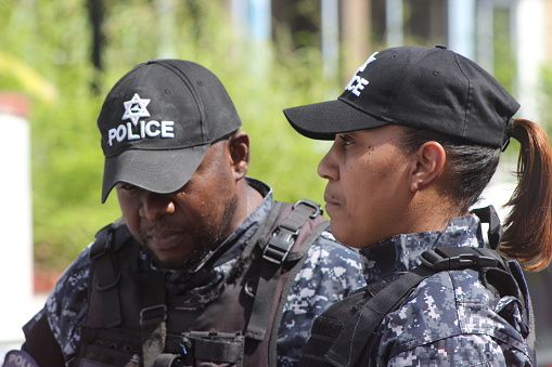 Port of Spain, Trinidad - August 31, 2022: \nCandid shot of Two young police officers strapped and dressed in their camouflaged uniforms, on duty for the 60th Independence Day parade