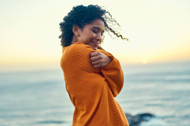 Happy black woman embrace herself on a beach at sunrise, self love and peaceful. Young African American feeling in touch with earth and nature, enjoy freedom and inner peace, smiling, loving soul stock photo