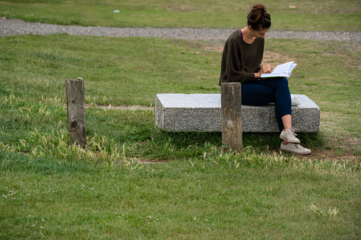 London, UK - June 2020 : Young woman sitting on a concrete slab bench and reading a book on a hot summer day