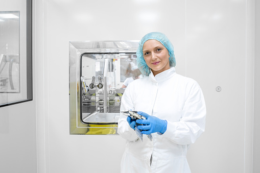 Portrait of a young smiling female employee in a pharmaceutical industry completely dressed in a protective workwear seen holding a few blister packs with white pills just after manufacturing process.