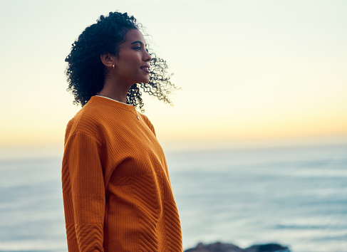 Woman at beach during sunset in nature, happy at sea in nature on holiday and content with sky at ocean during travel vacation. African person with smile in summer by calm water with sun in horizon