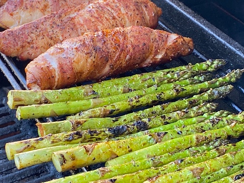 Image of bacon wrapped chicken and fresh asparagus on the top rack of a bbq being grilled for dinner