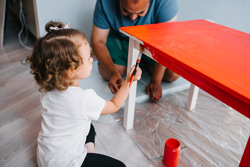 Little preschool age girl painting table in red color with her father. Furniture repair.