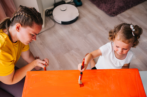 Mother with preschool age daughter painting table in red color. Furniture repair.