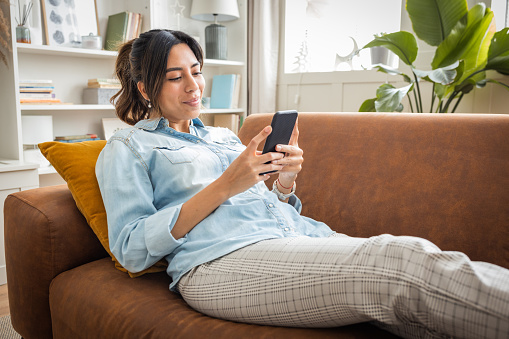 Smiling woman using mobile phone while sitting on sofa. She is text messaging in living room. She is relaxing at home.