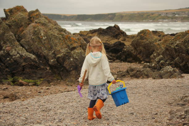 Little girl walking along pebble beach carrying bucket and spade towards sea